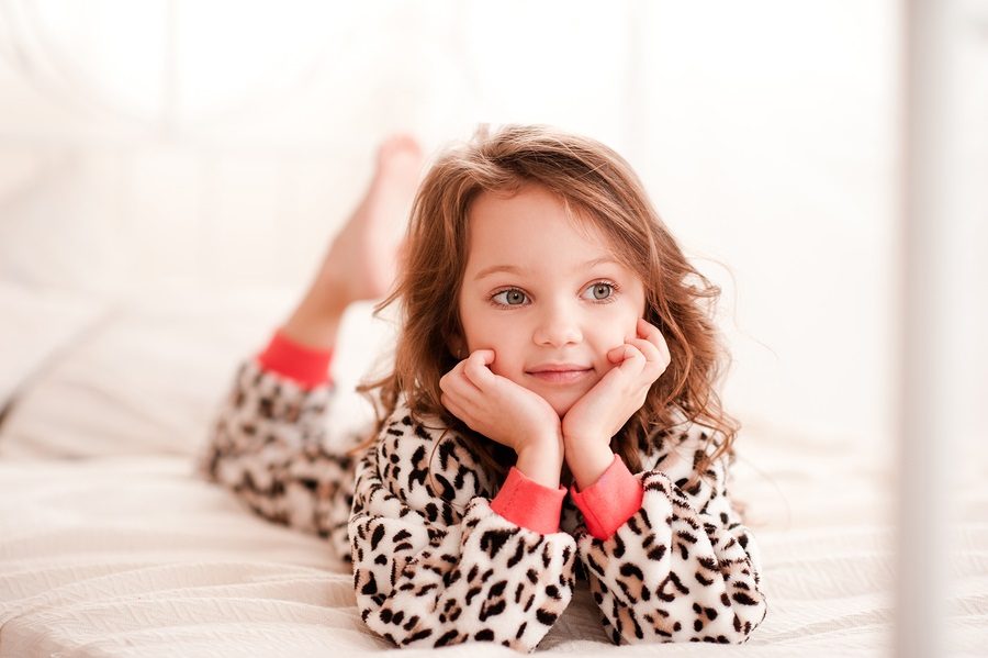 Smiling kid girl 5-6 year old lying in bed wearing leopard print pajamas in room. Looking away. Childhood. Resting in bed. Bed time.
