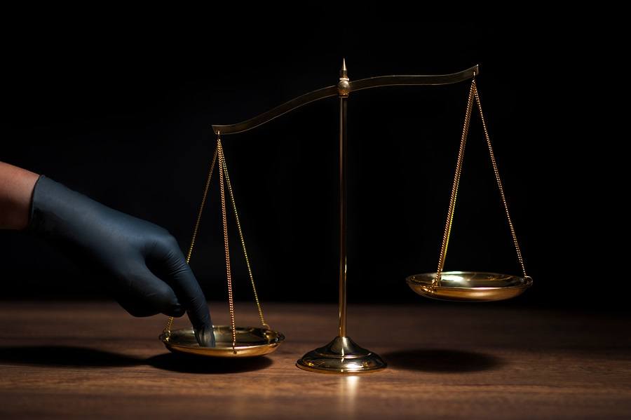 Hand with black glove pushing on scale of justice in dark room with black background. 